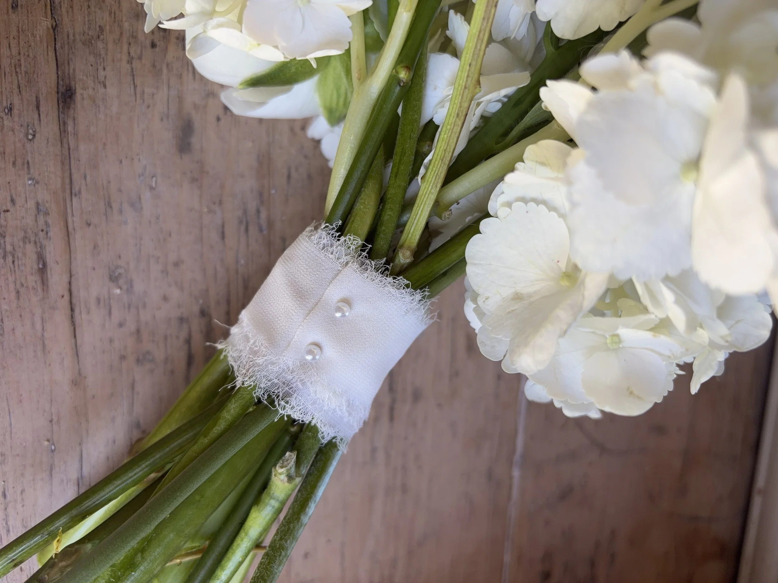 White hydrangea flowers with green stems wrapped in white fabric with pearl embellishments on a wooden surface.