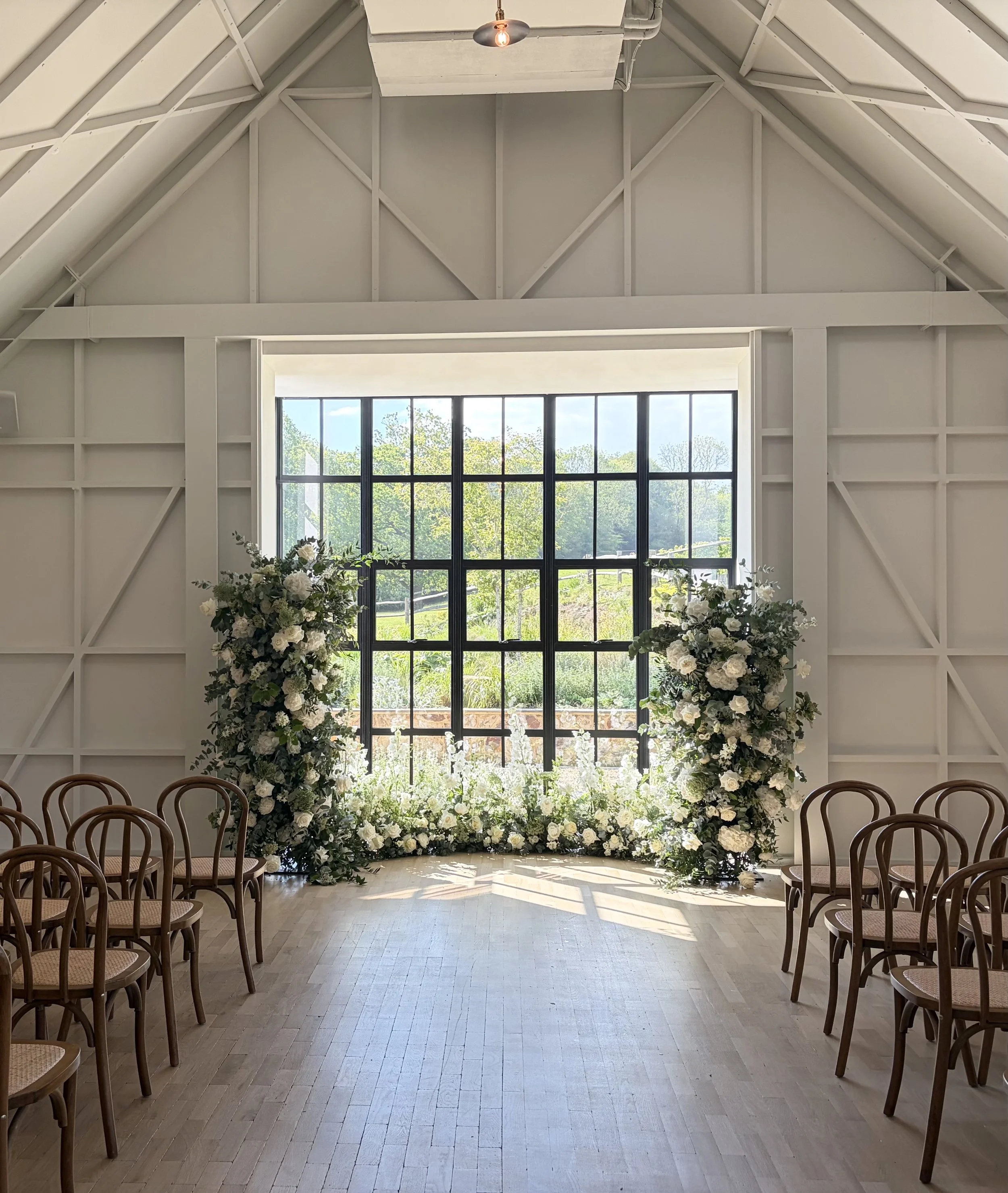 Interior of a modern venue with high ceiling, large window, arranged chairs for a ceremony, floral arrangements on either side of a floral archway, and natural light streaming in.