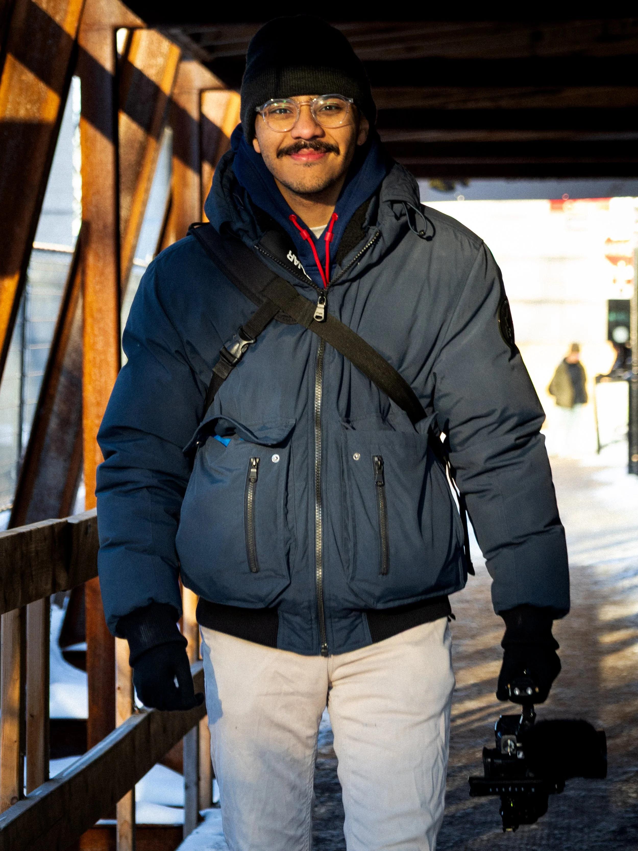 Un homme souriant portant une veste d'hiver, une casquette noire, des lunettes, et des gants, avec un snowboard à ses pieds, dans un environnement enneigé.