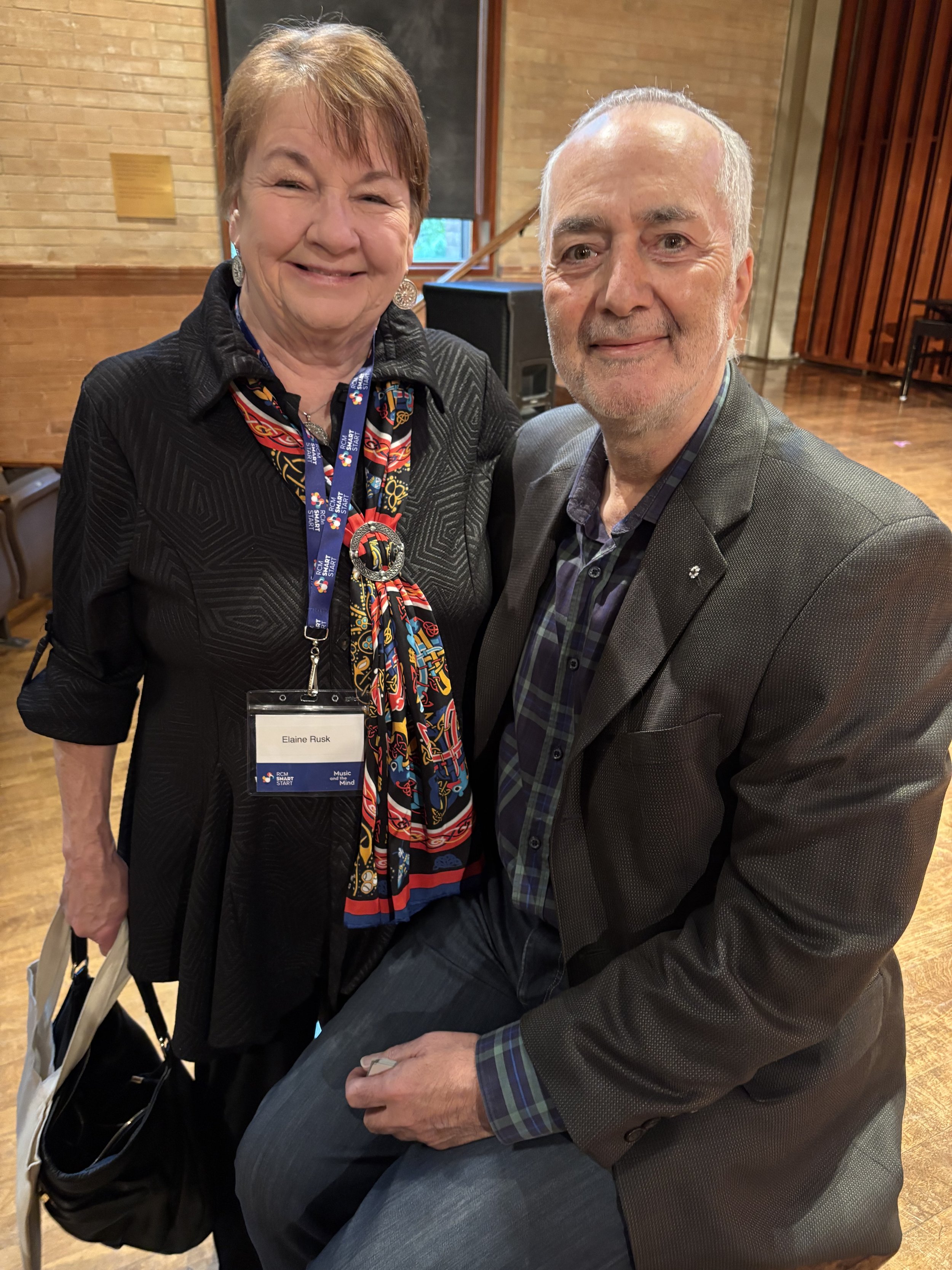 An elderly woman and a man pose together in an indoor setting. The woman is wearing a black jacket, colorful scarf, and name tag, and is smiling. The man is wearing a gray blazer, plaid shirt, and jeans, and is also smiling. The woman has a bag in her hand, and there is a brick wall and wooden floor in the background.