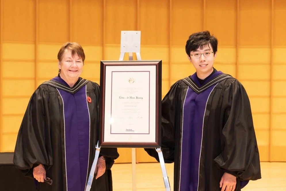 A woman and a young man in academic regalia standing next to a framed certificate and a small easel in a room with wood-paneled walls.