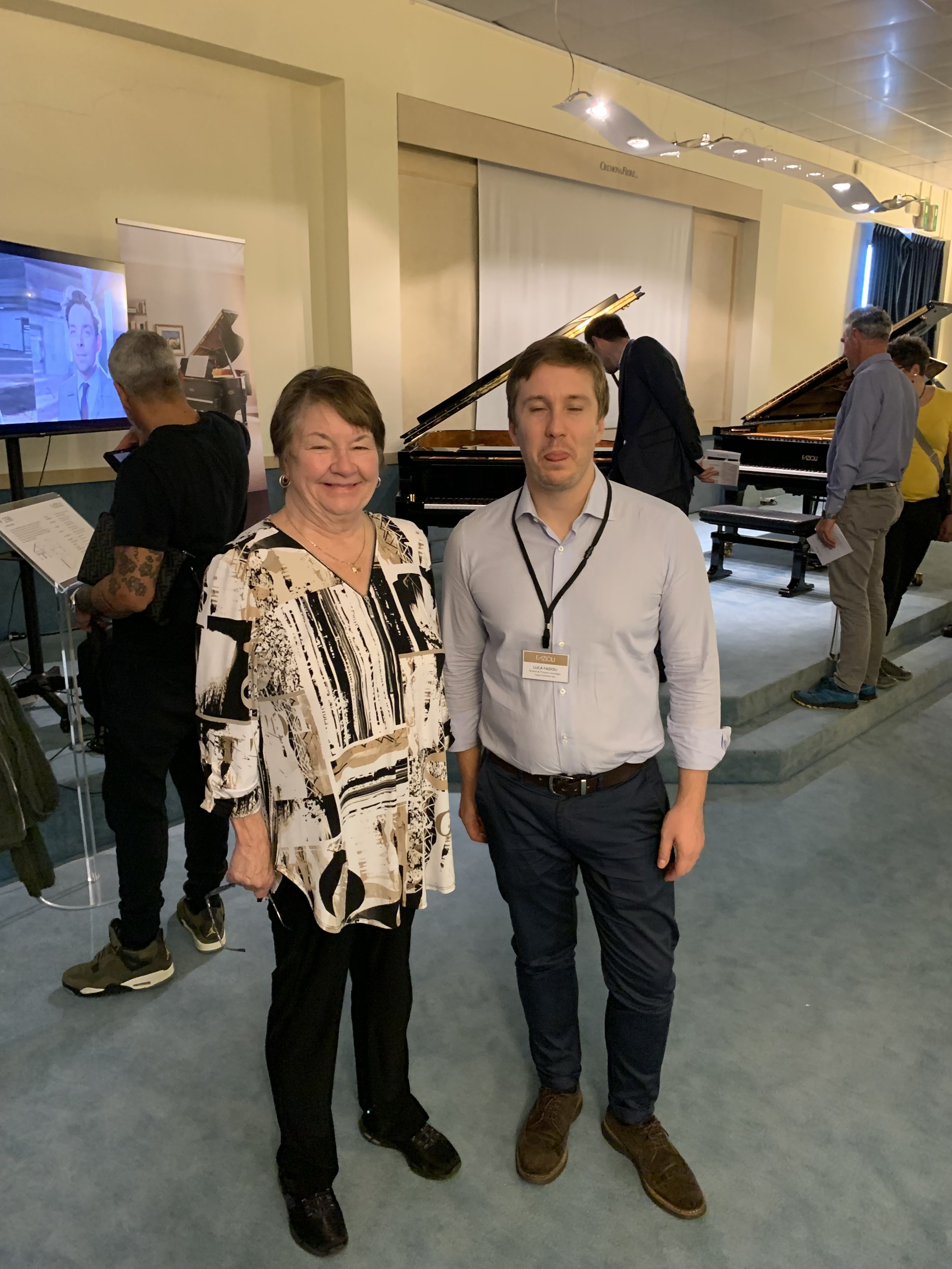 A photograph of two people standing together in a room with several pianos. The woman on the left is smiling and wearing a patterned blouse and black pants. The man on the right is making a funny face, wearing a light-colored shirt and dark pants, with a lanyard around his neck. Other people are in the background, some looking at the pianos and a television screen.
