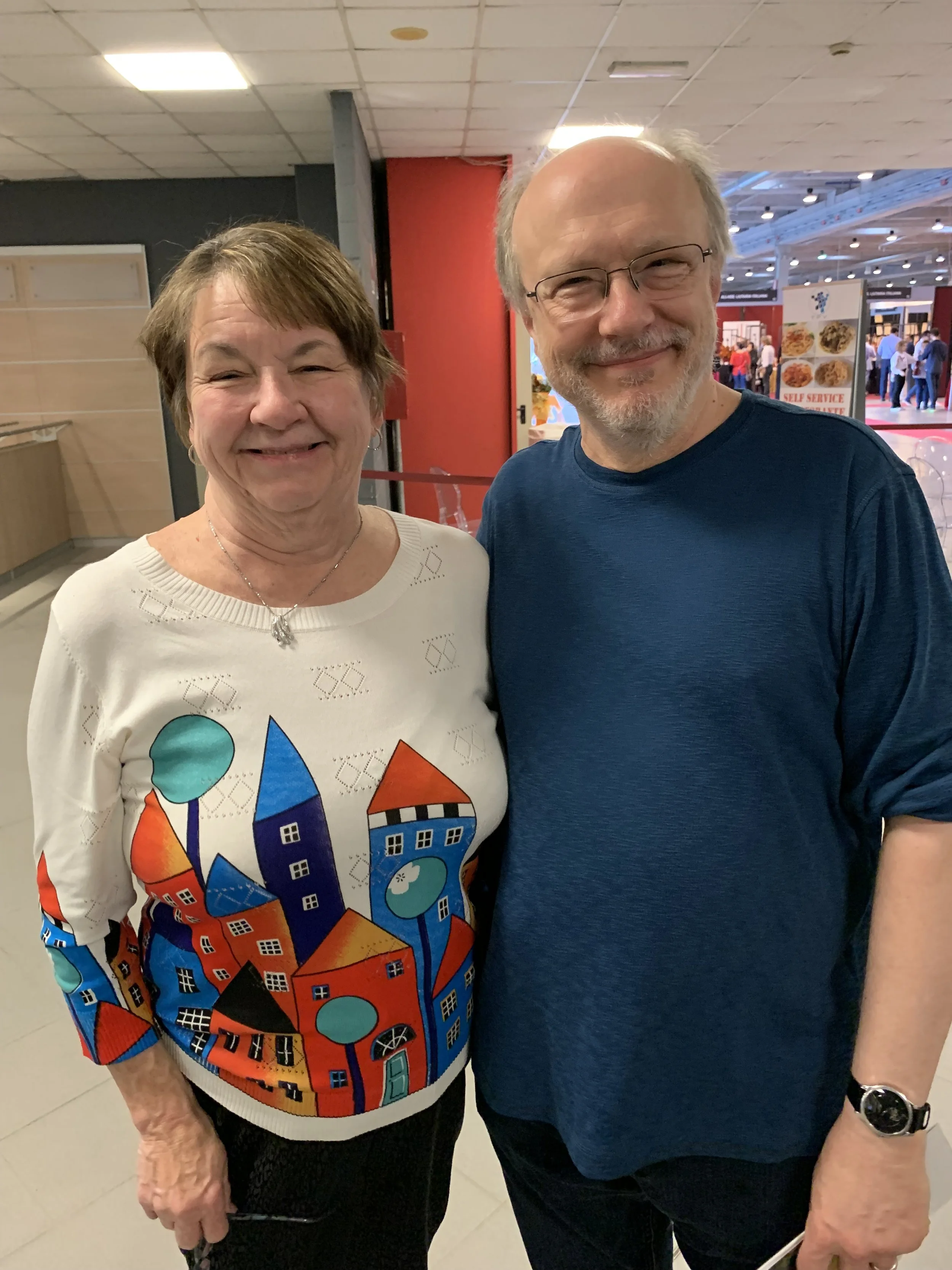 A smiling elderly woman in a colorful sweater with house and tree designs standing next to a smiling man in a blue shirt, in an indoor public space with food stalls visible in the background.