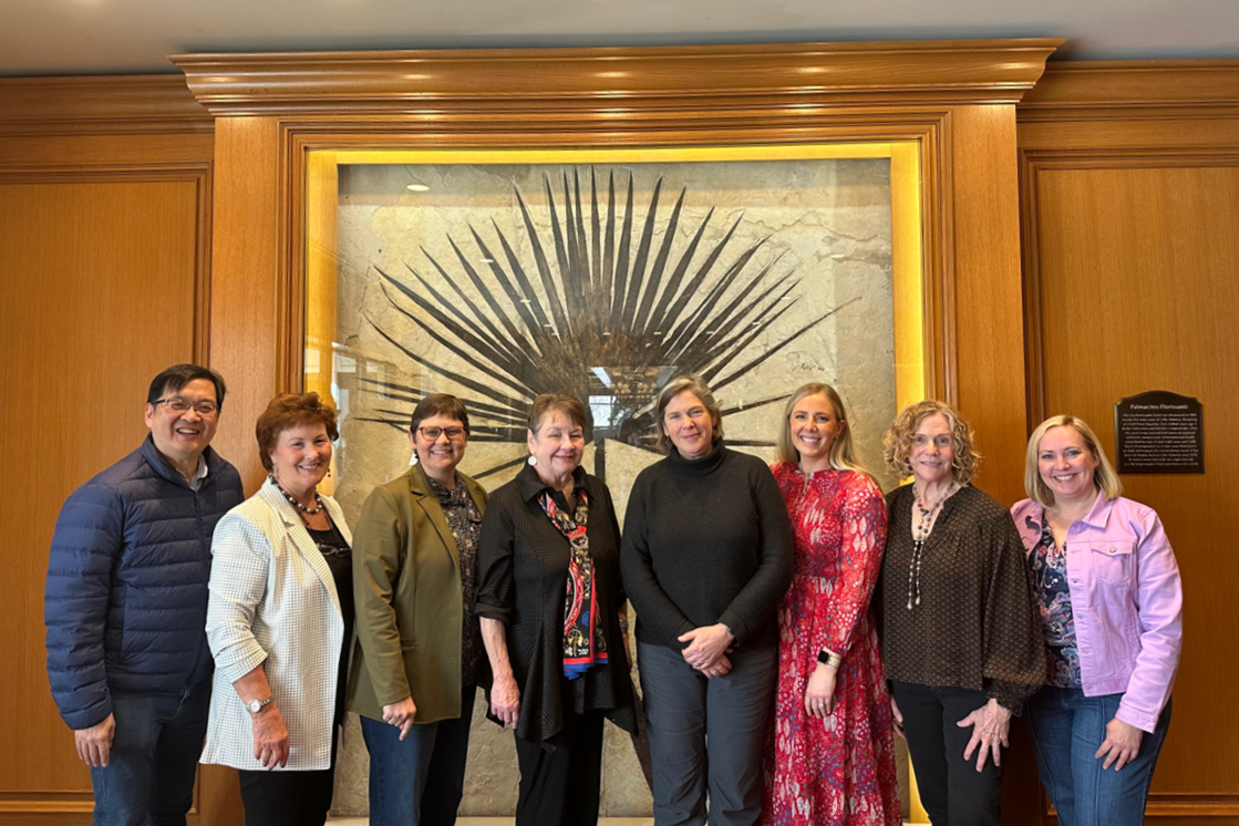 Group of eight diverse women standing in front of a large framed fossil display on wooden wall, smiling at camera.