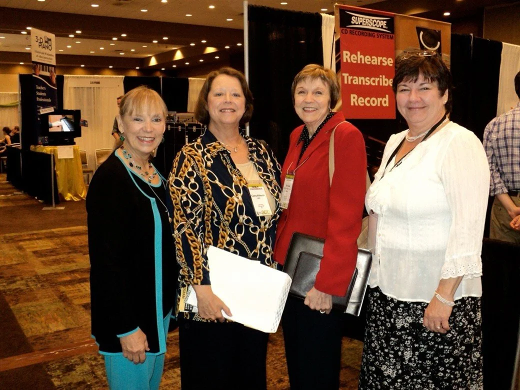 Four women standing together at a conference, smiling, with a booth behind them promoting a CD recording system.