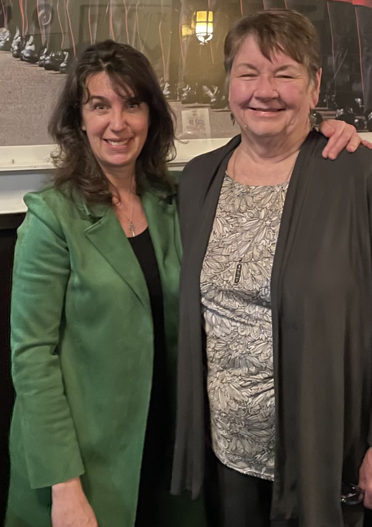 Two women standing close together, smiling, in an indoor setting with a background featuring a banner and wall decor.