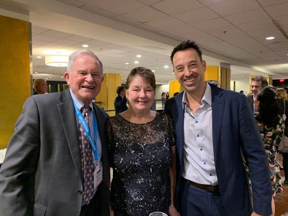Three smiling people, two men and one woman, dressed in formal attire, standing together at an indoor event.