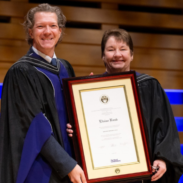 Two people in academic robes holding a framed certificate during a graduation or award ceremony.