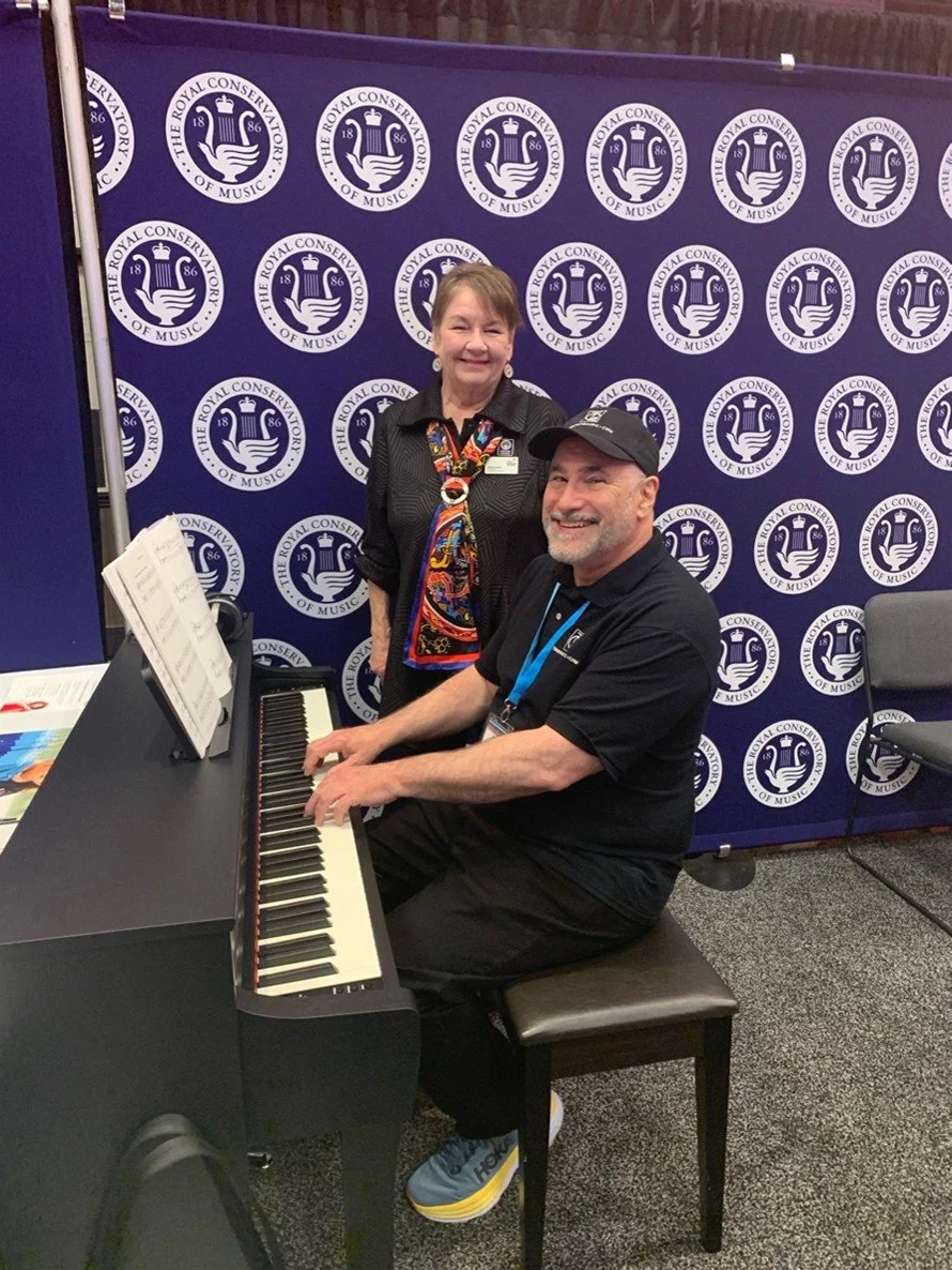 A woman and a man are at a music event in front of a backdrop with 'The Royal Conservatory of Music' logos. The man is sitting at a black piano, wearing a black t-shirt, cap, and sneakers, smiling and playing the piano. The woman is standing behind him, wearing a black blouse and a colorful scarf, smiling at the camera.