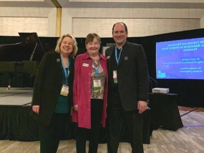 Three people standing together at a conference, smiling. The background includes a stage with a screen and a piano.