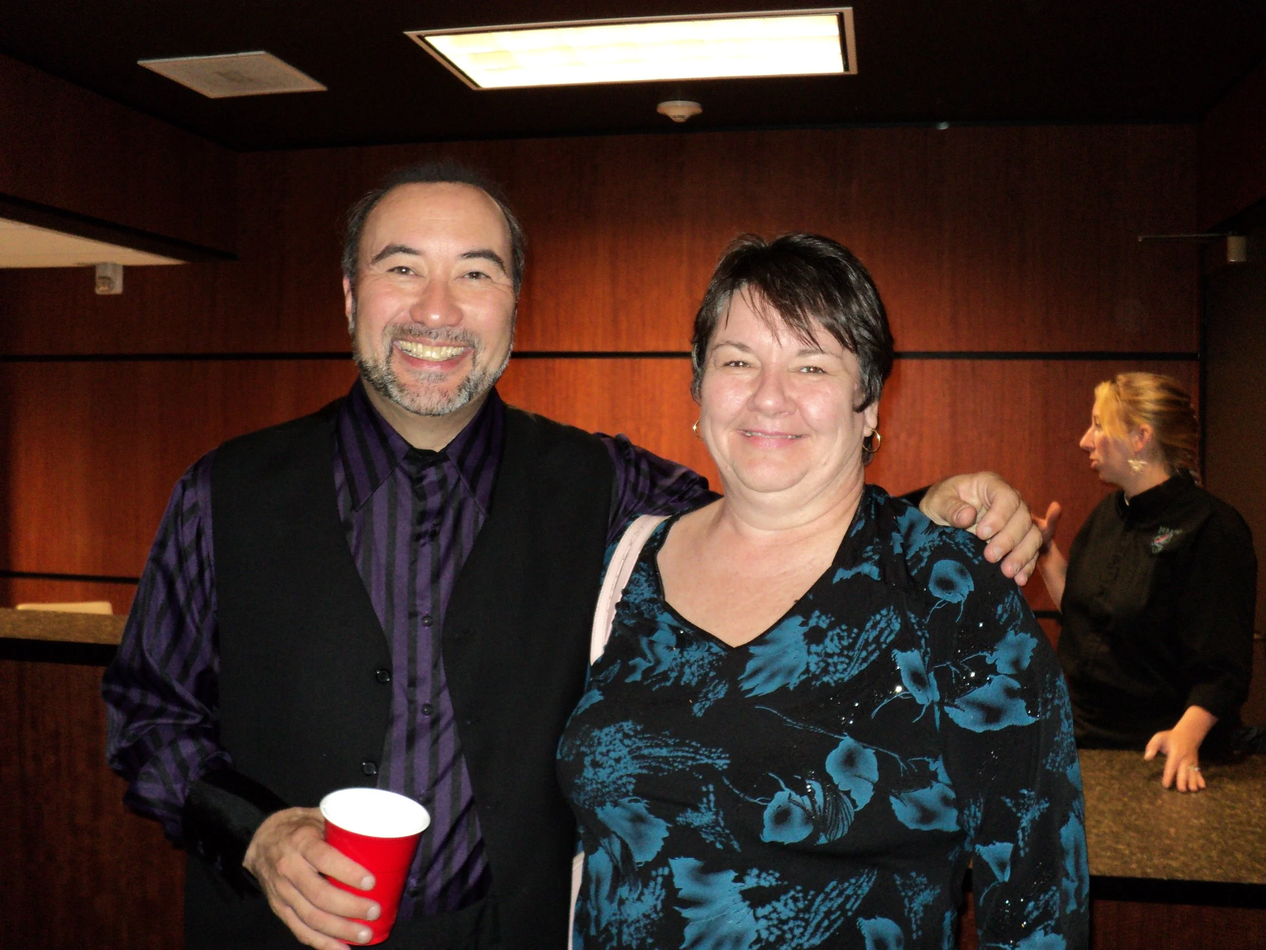 A smiling man holding a red cup and a woman smiling, standing together in a room with wood-paneled walls, with another woman talking in the background.