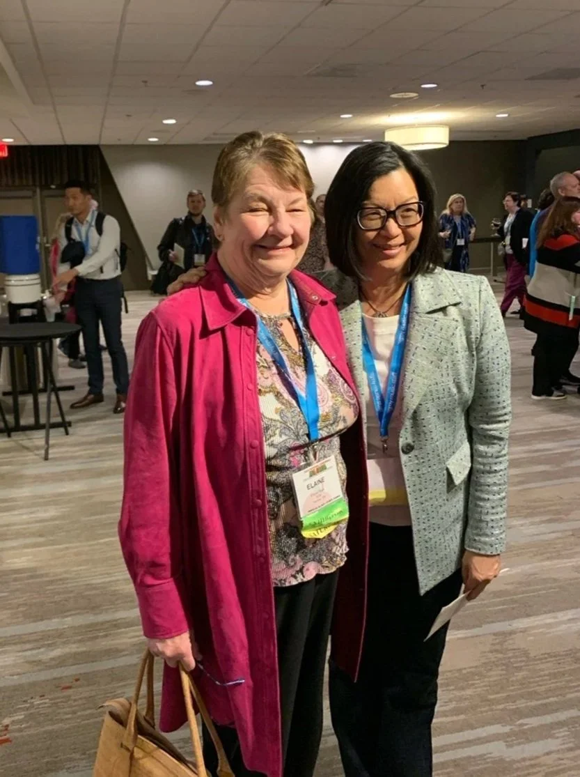 Two women smiling at a conference, standing close together, wearing conference badges, in a crowded indoor hallway or event space.