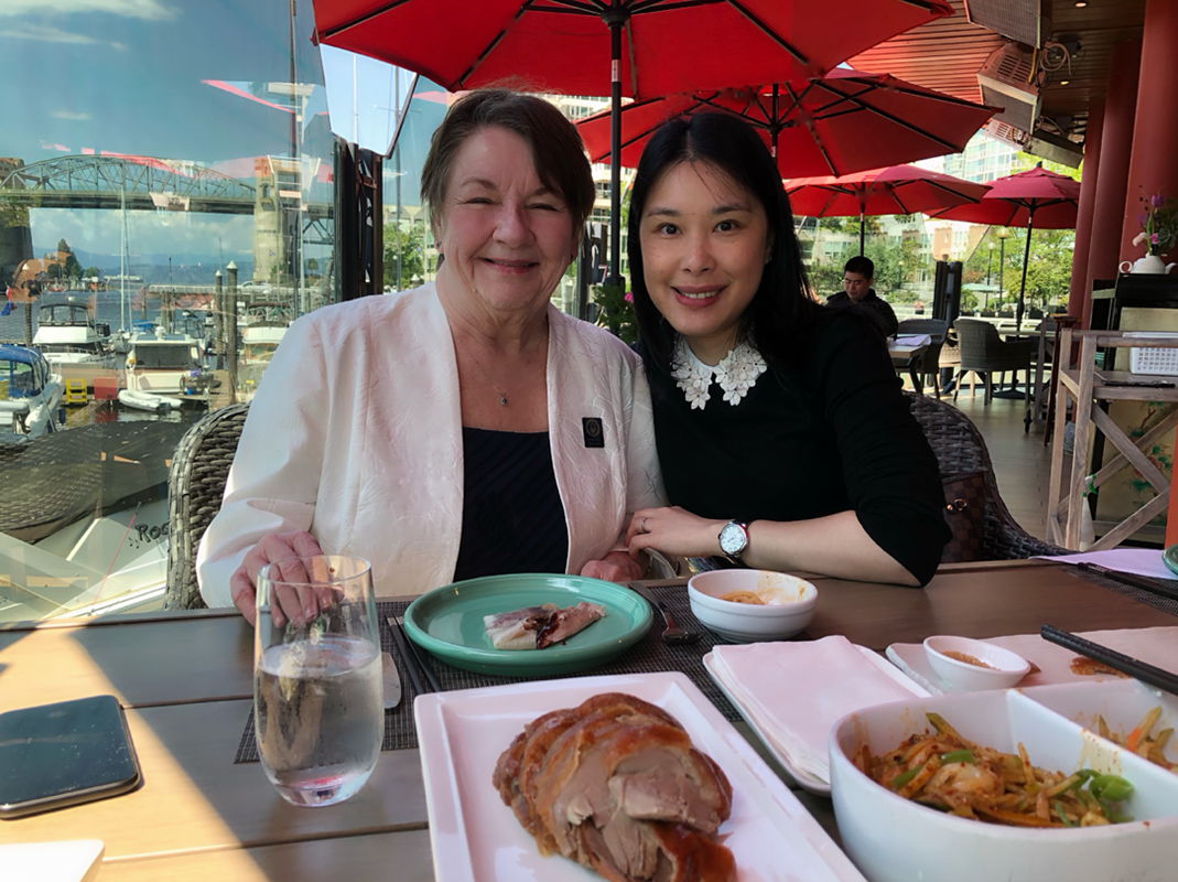 Two women sitting at an outdoor restaurant table during daytime, smiling. The table has Asian dishes, including sliced meat, spring rolls, and vegetables. Red umbrellas are overhead; a harbor with boats and a bridge is visible in the background.