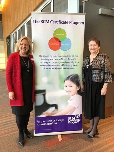 Two women standing next to a poster for The RCM Certificate Program, which offers comprehensive music study and assessment based in North America, featuring a smiling young girl on the poster.