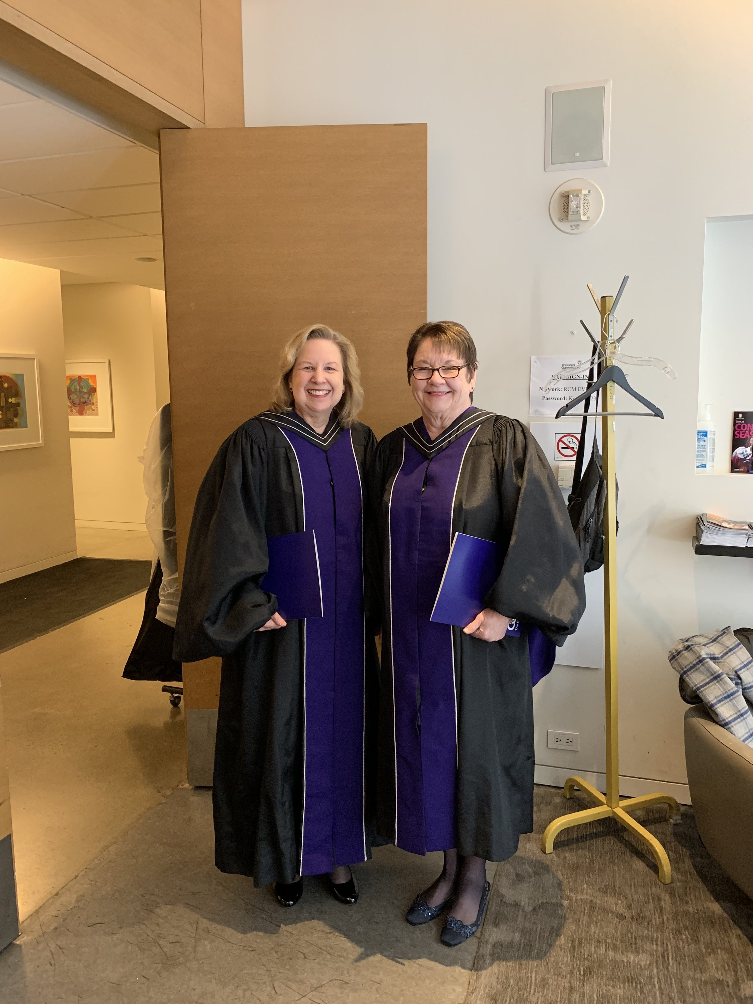 Two women in graduation gowns and caps standing indoors, smiling, holding diplomas, in front of a wooden partition and a wall with some artwork, coat rack, and signs.