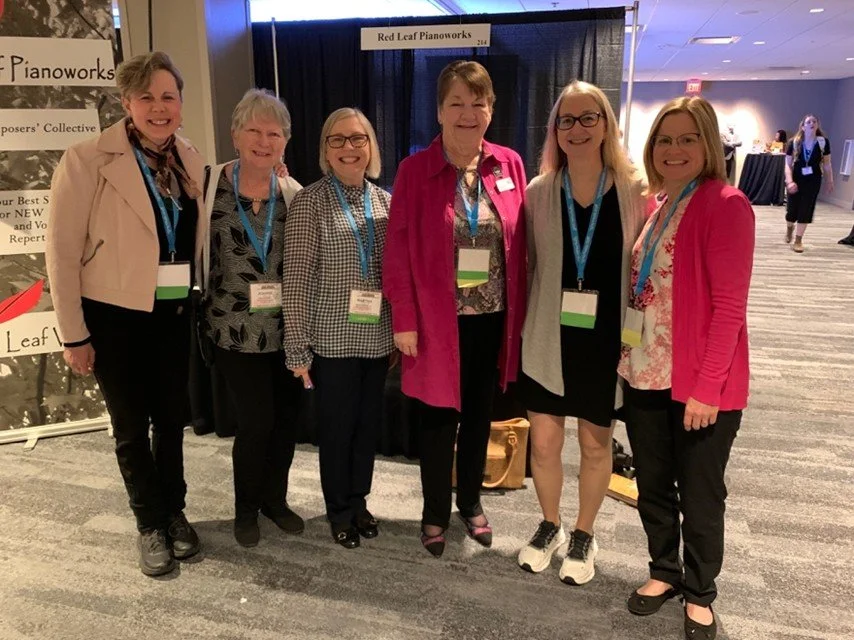 Six women standing together at a conference, smiling, posing for a photo indoors with a black curtain backdrop, some wearing name tags and lanyards.