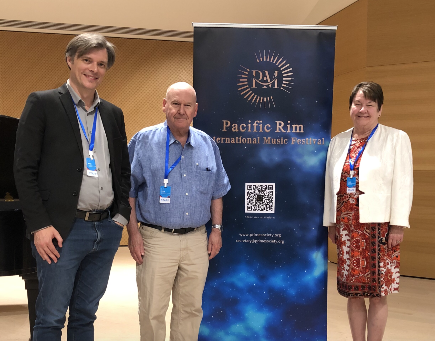 Three people standing in front of a banner at the Pacific Rim International Music Festival, with a grand piano visible on the left.