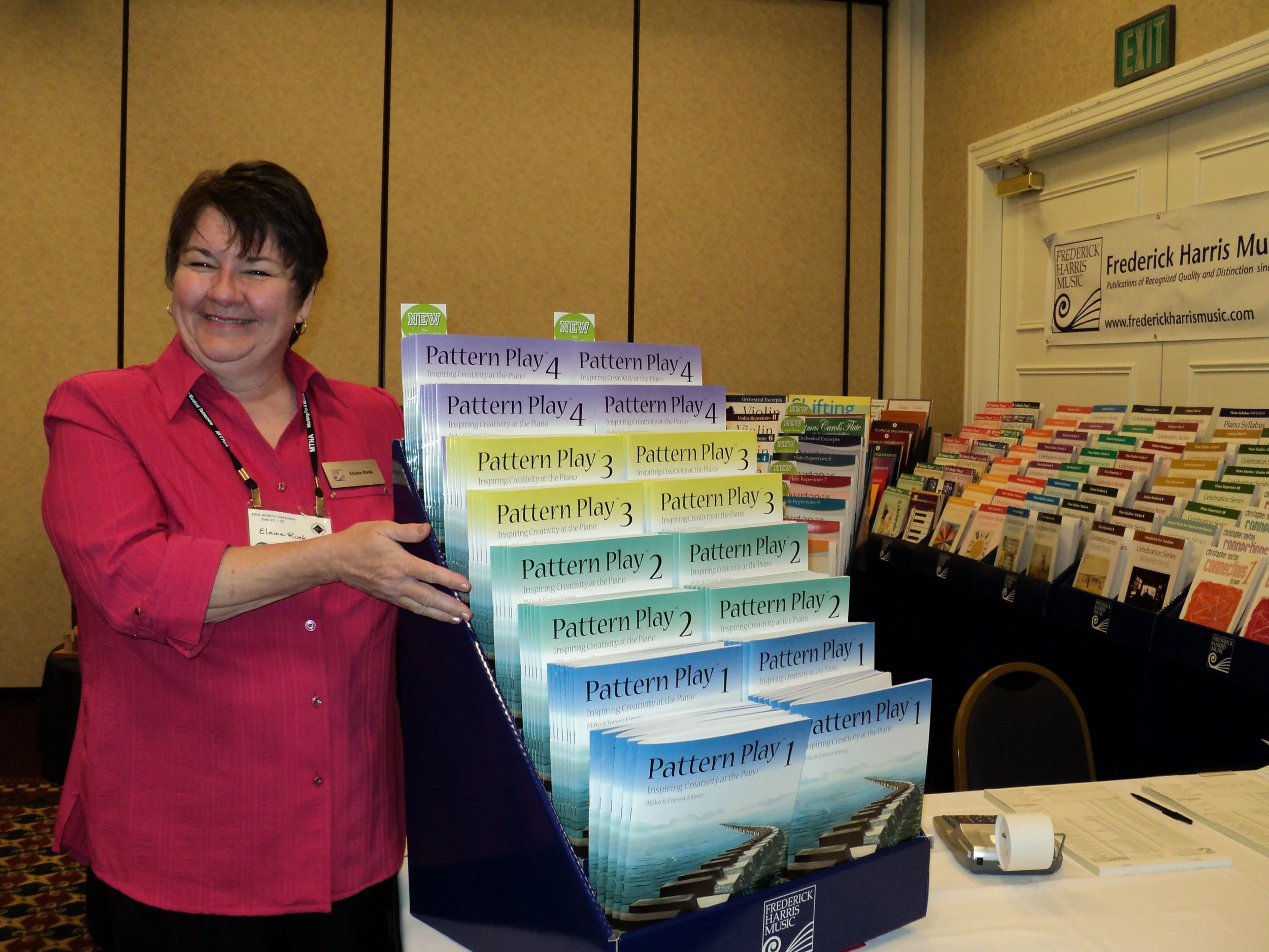 A woman in a red shirt holds a display of music books titled 'Pattern Play' at a conference booth, with a table, stack of papers, and a banner in the background.