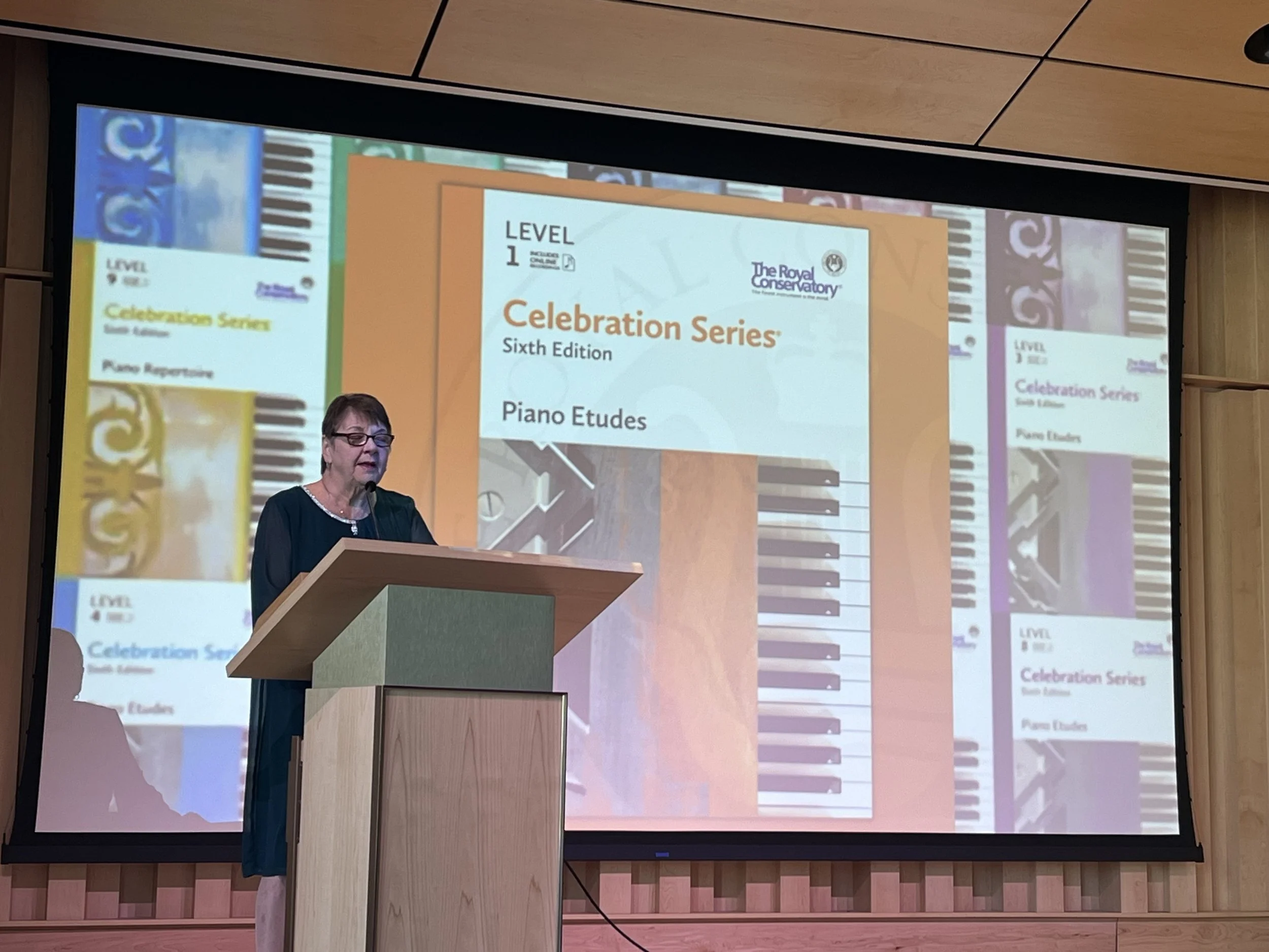 A woman with glasses and short hair is standing at a podium, giving a presentation in front of a large screen displaying a slide titled 'Celebration Series, Sixth Edition, Piano Etudes.'