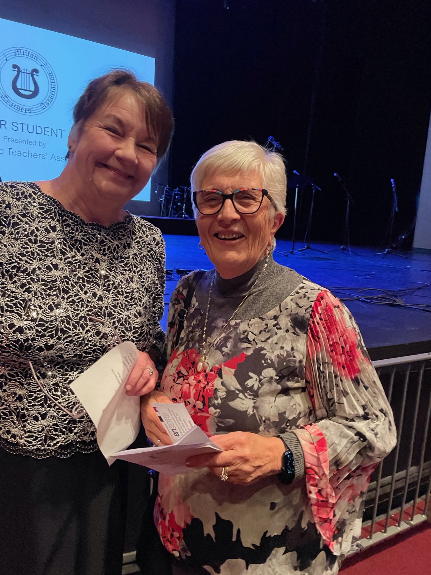 Two smiling women standing close together, holding a piece of paper and a ticket, at an indoor event with a stage in the background.