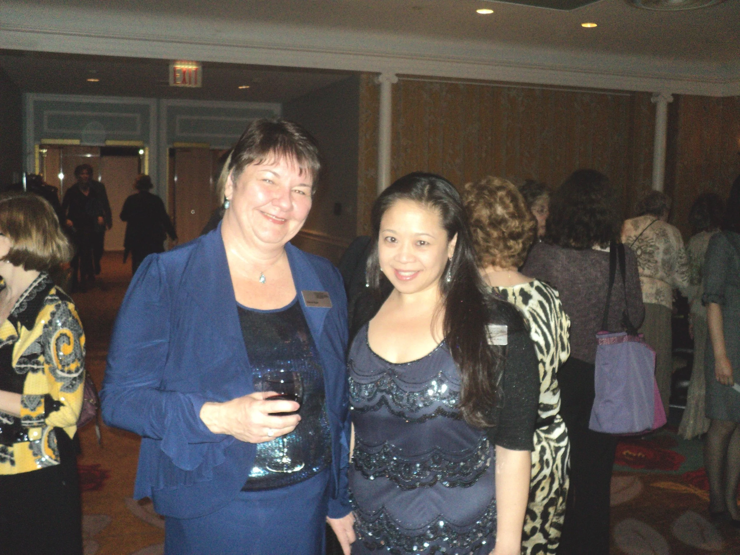 Two women at a social event, one holding a glass of red wine, with others in the background.