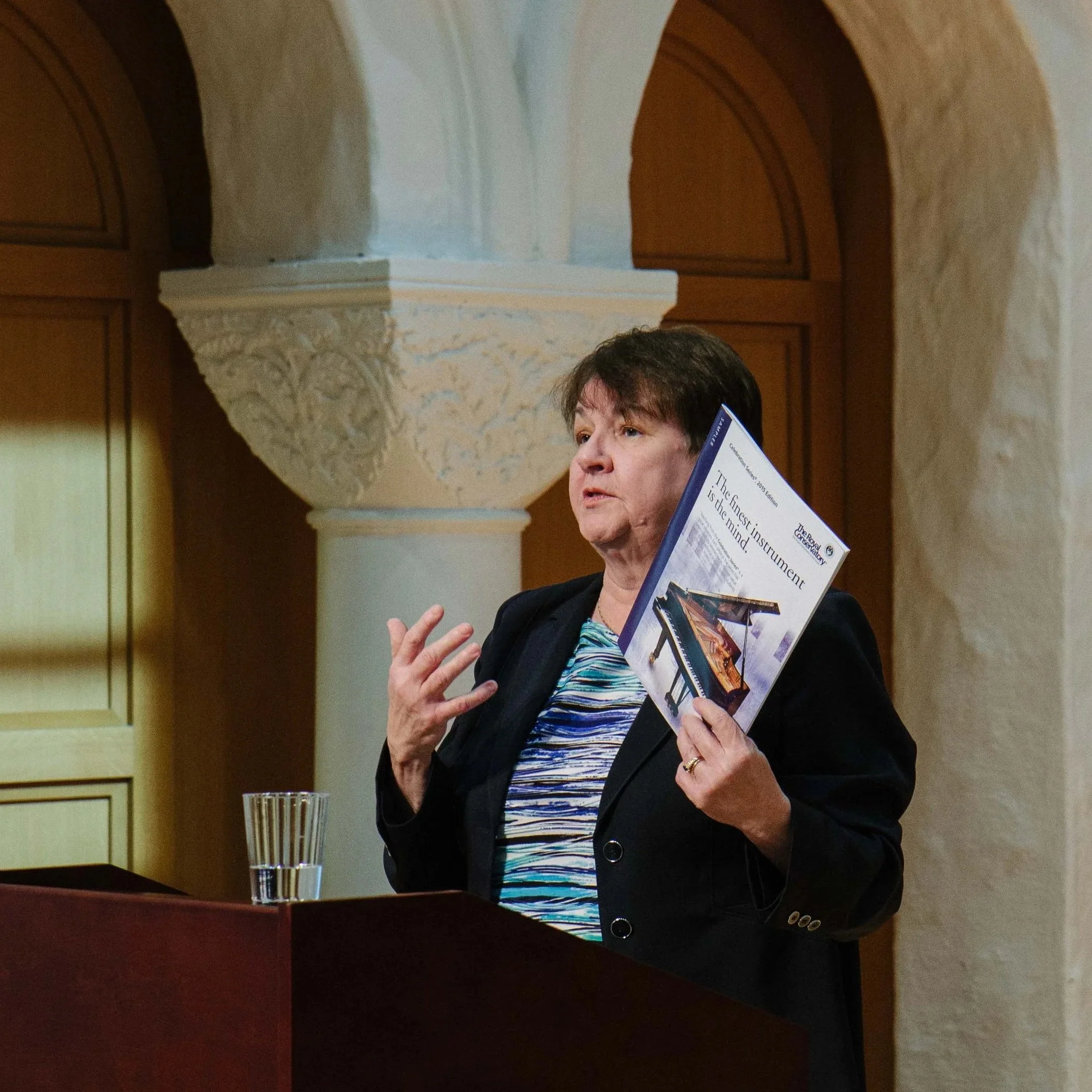A woman with short dark hair wearing a black blazer and a striped shirt holding a brochure about a grand piano, standing at a podium with a glass of water, inside an ornate room with cream-colored walls and decorative columns.
