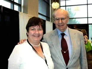 A woman and an elderly man smiling together indoors, with large windows behind them and other people in the background.