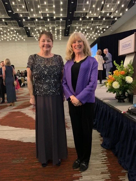 Two women standing together at an indoor event, smiling, with a stage and floral arrangement in the background.