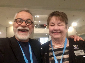 A smiling man with glasses and a beard, and a smiling woman with short hair, standing together at an indoor event.