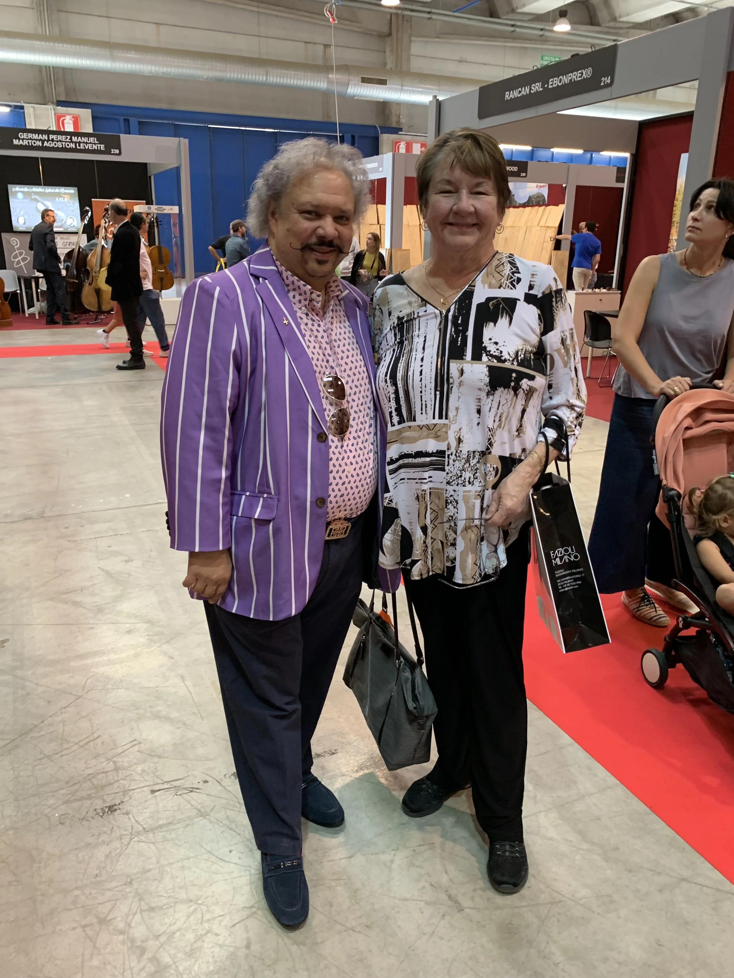 Two people posing together at an indoor event, one man wearing a purple striped blazer and a woman in a patterned blouse, surrounded by booths and other attendees.