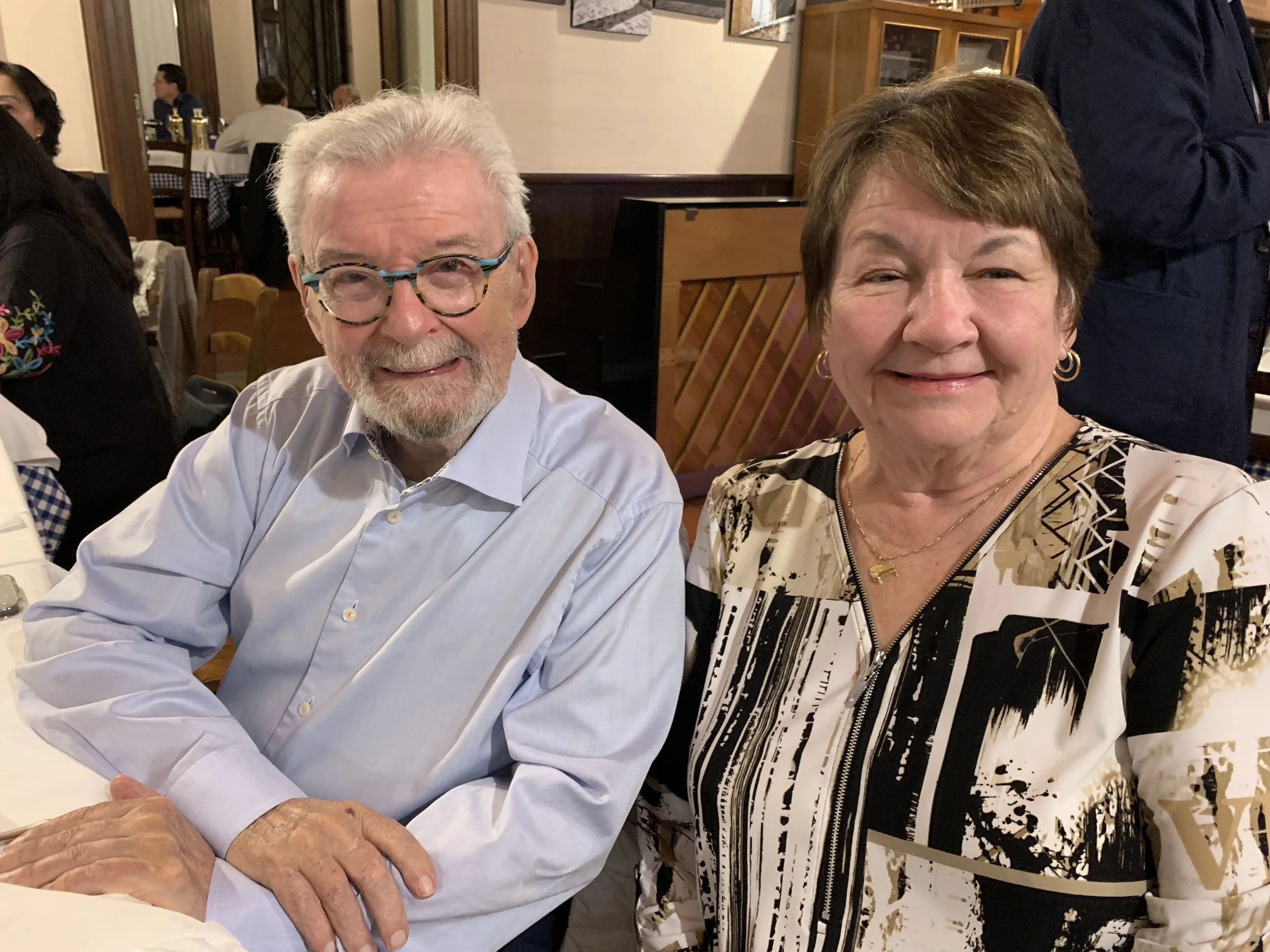 An elderly man and woman smiling at a restaurant; the man has white hair and beard, glasses, and is wearing a light blue shirt, while the woman has short brown hair, hoop earrings, and a patterned black and white blouse.