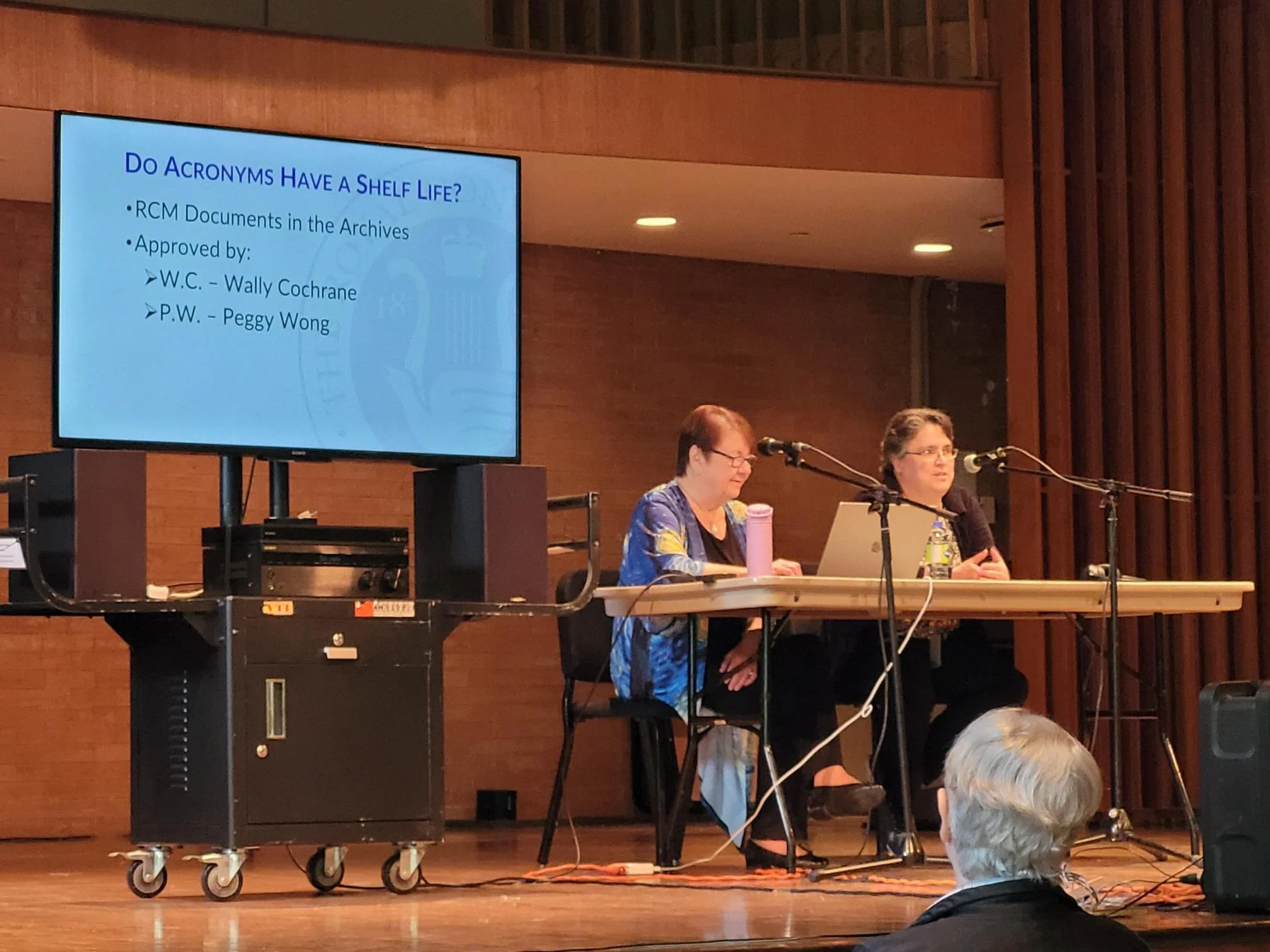 A presentation slide on a large screen displays the title 'Do Acronyms Have a Shelf Life?' with bullet points about RCM documents and approval by W.C. Wally Cochrane and P.W. Peggy Wong. Two women sit at a desk on a stage with microphones, one of whom appears to be speaking into a microphone, while the other looks at a laptop. An audience member with gray hair is visible in the foreground.