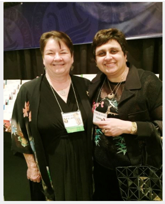 Two women standing close together, smiling at a conference or event, with name tags around their necks.