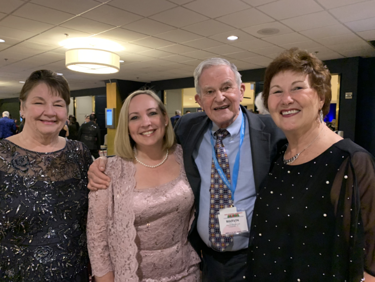 Four adults, three women and one man, smiling and posing closely together at a social event in an indoor venue.