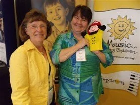 Two women smiling and holding a puppet, standing in front of a banner with the word 'Music' and a sun illustration.