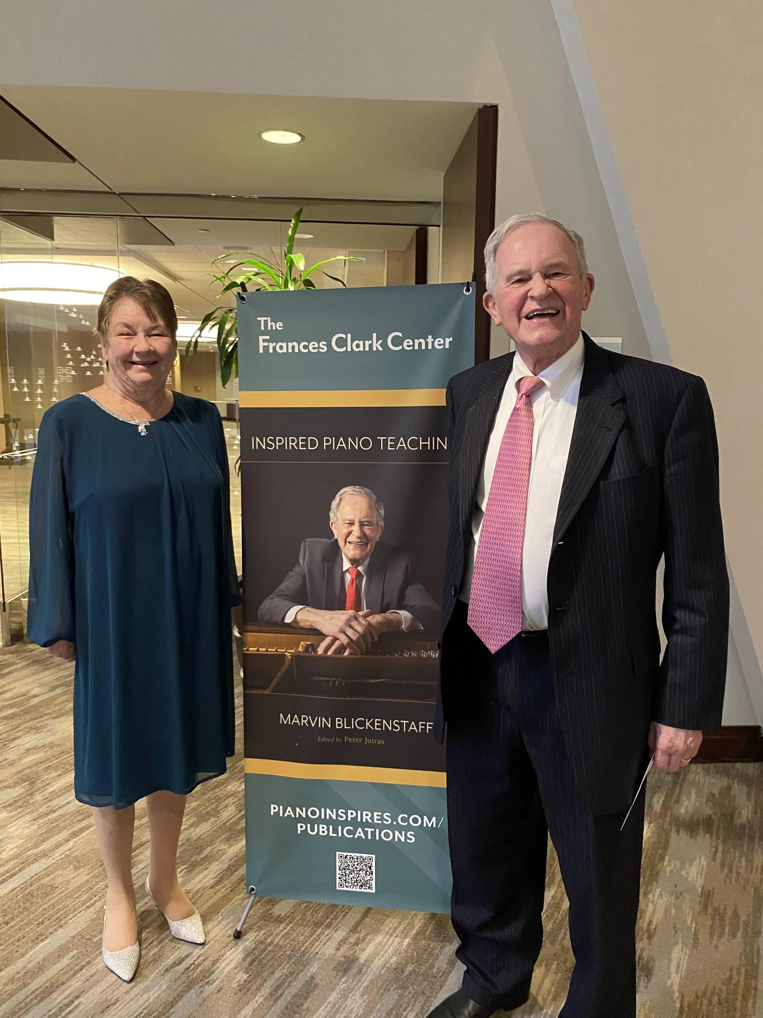 Two smiling elderly individuals standing beside a vertical banner for The Frances Clark Center, which features a photograph of a man at a piano and text about inspired piano teaching by Marvin Blickenstaff. They are indoors on a carpeted floor with a potted plant in the background.