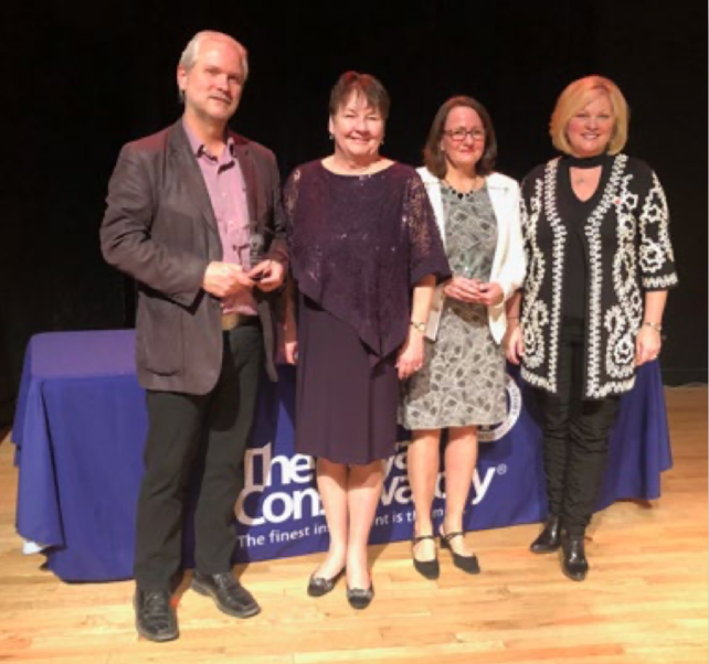 Four people standing on a stage in front of a table with a blue tablecloth and a banner that reads 'The Company of...' with the rest obscured, at an indoor event.