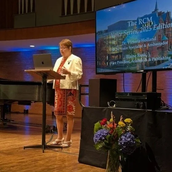 A woman standing at a podium giving a presentation in a room with a large screen displaying the title 'The RCM Voice Series, 2025 Edition' and details about speakers, with a floral arrangement in the foreground.