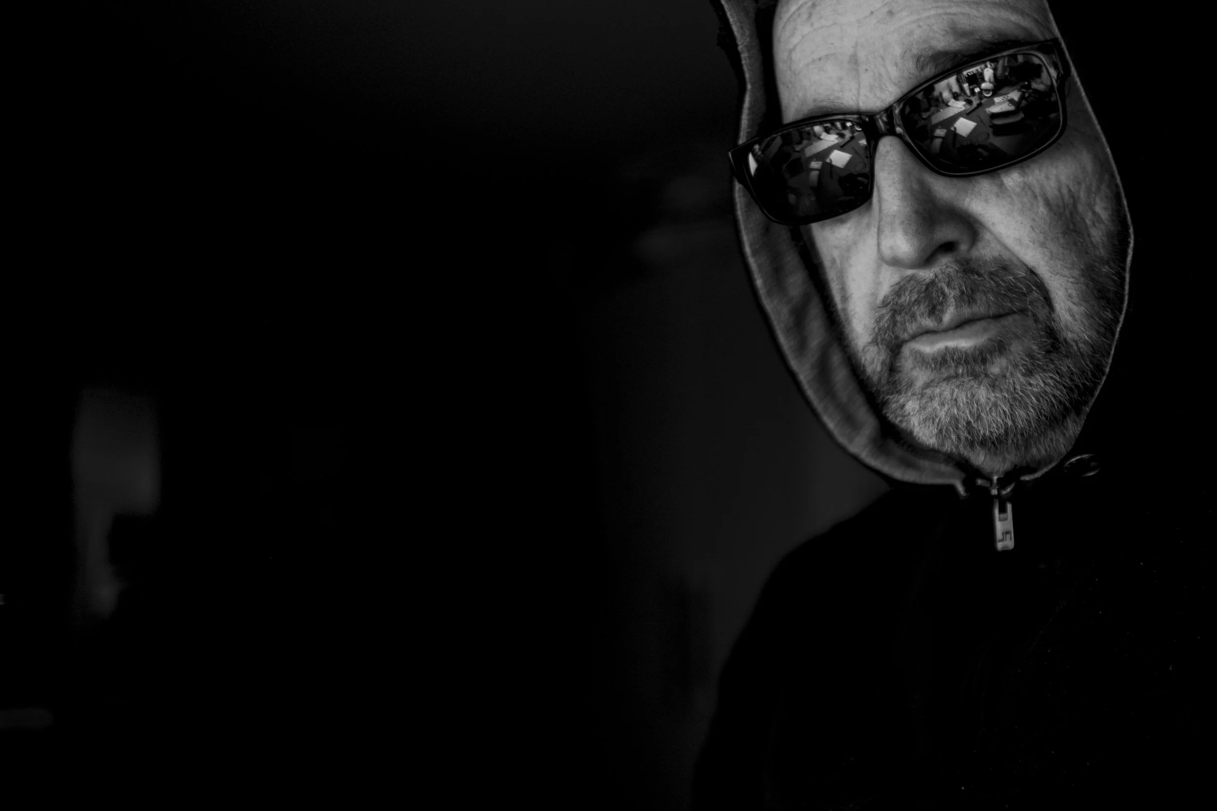Schwarz-weiß-Foto eines Mannes mit Bart und Sonnenbrille in dunkler Kleidung, nur die rechte Gesichtshälfte sichtbar.