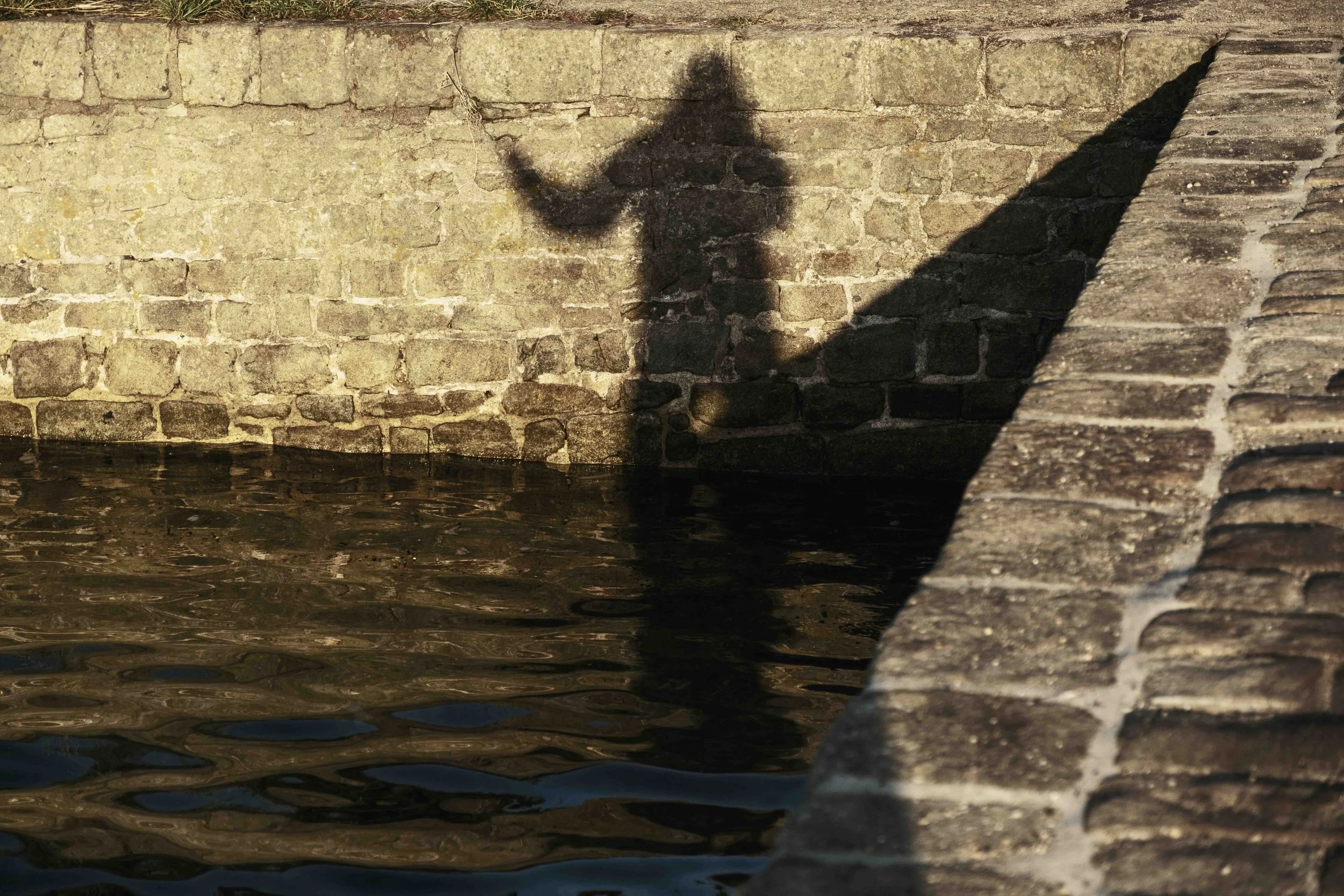 Schatten einer Person, die eine Hand in die Luft hebt, auf einer Steinmauer an einem Wasserbecken bei Sonnenuntergang, mit Wasser im Vordergrund.