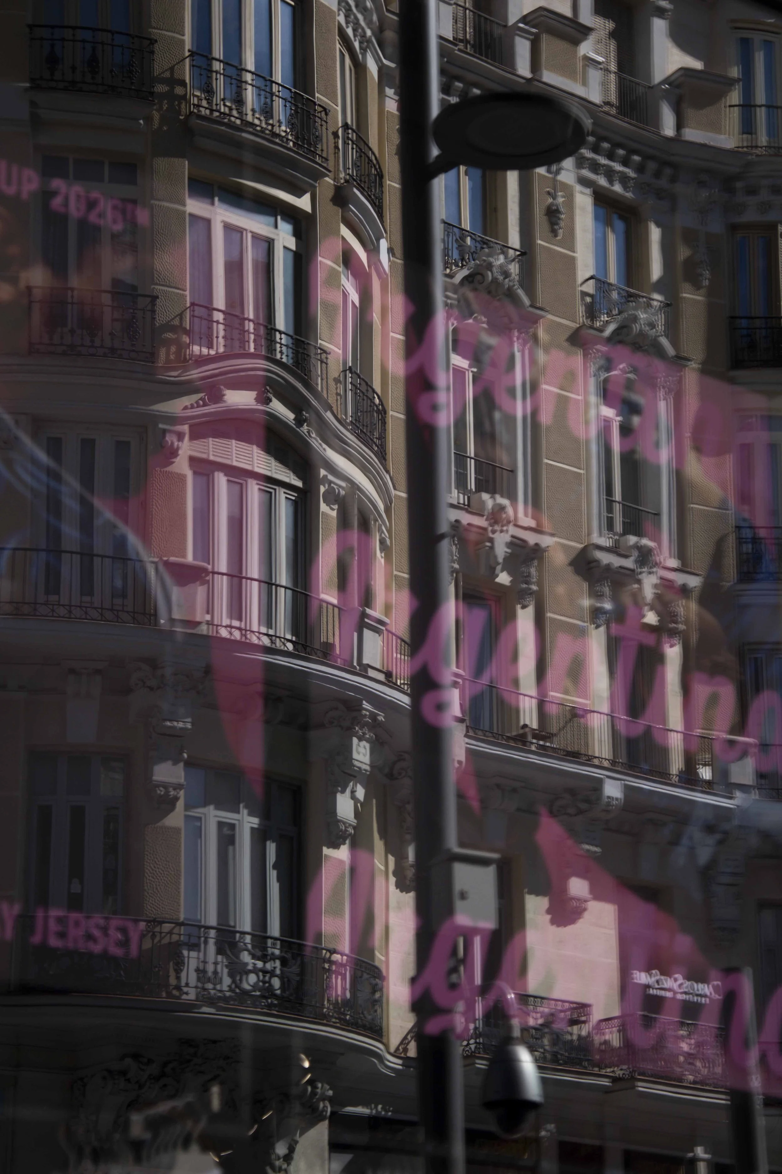 Reflexion eines historischen Gebäudes mit Balkon und Verzierungen in einem Fenster oder einer Glasscheibe, mit pinkem Text im Vordergrund.