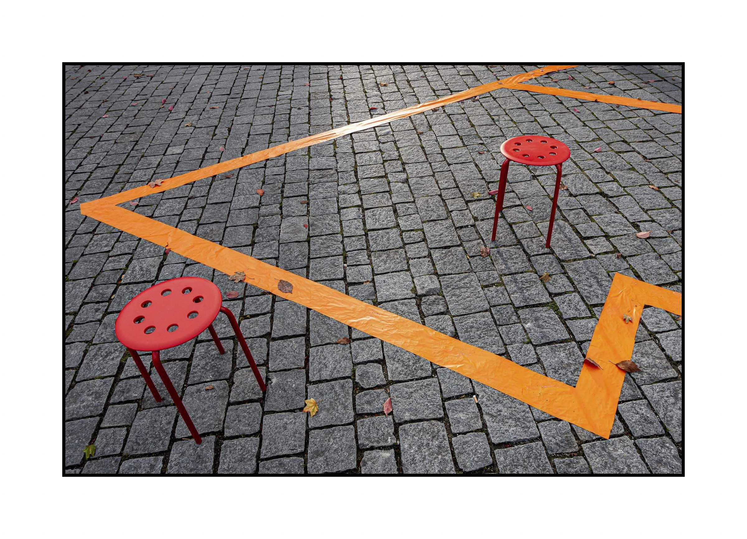 Aufgedruckte orangefarbene Absperrbänder auf einem gepflasterten Boden, umgeben von zwei roten Hockern.