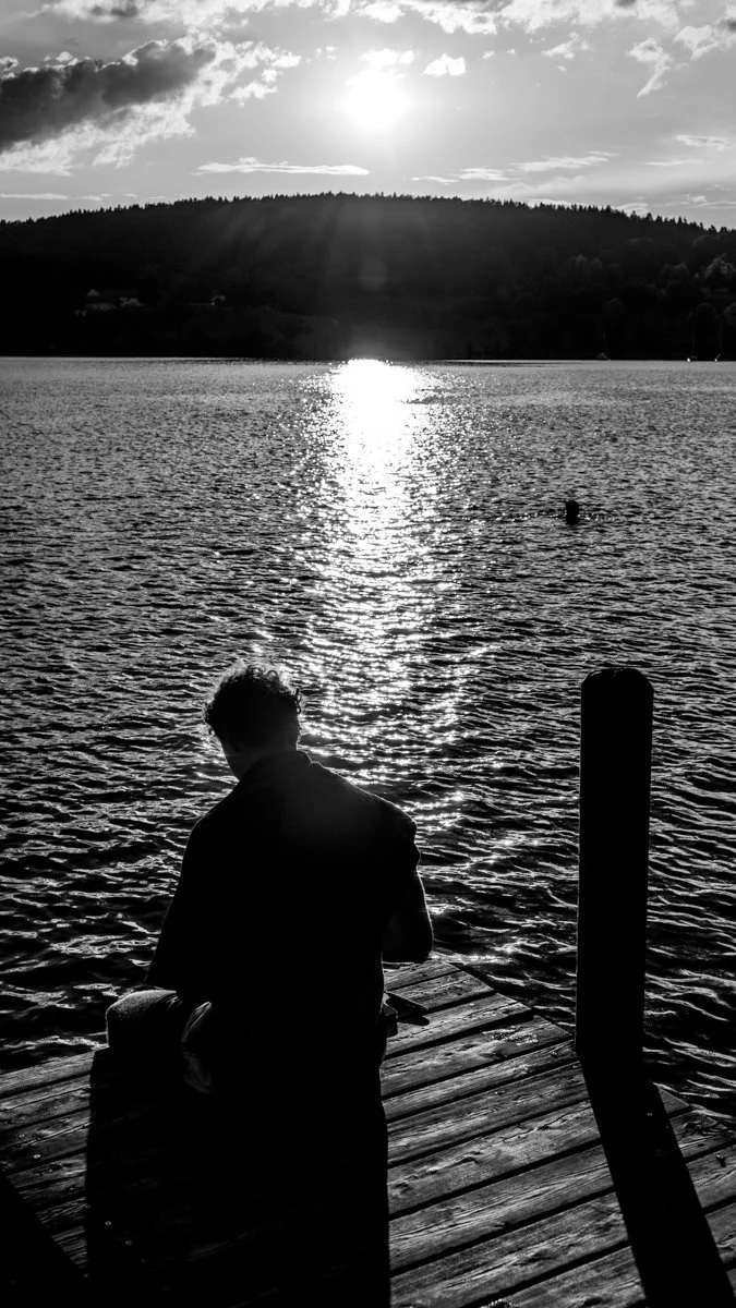 Eine silhouettierte Person sitzt an einem Steg am Wasser, blickt auf den Sonnenuntergang über einem See mit bewaldeten Hügel im Hintergrund. Die Szene ist in Schwarz-Weiß gehalten.