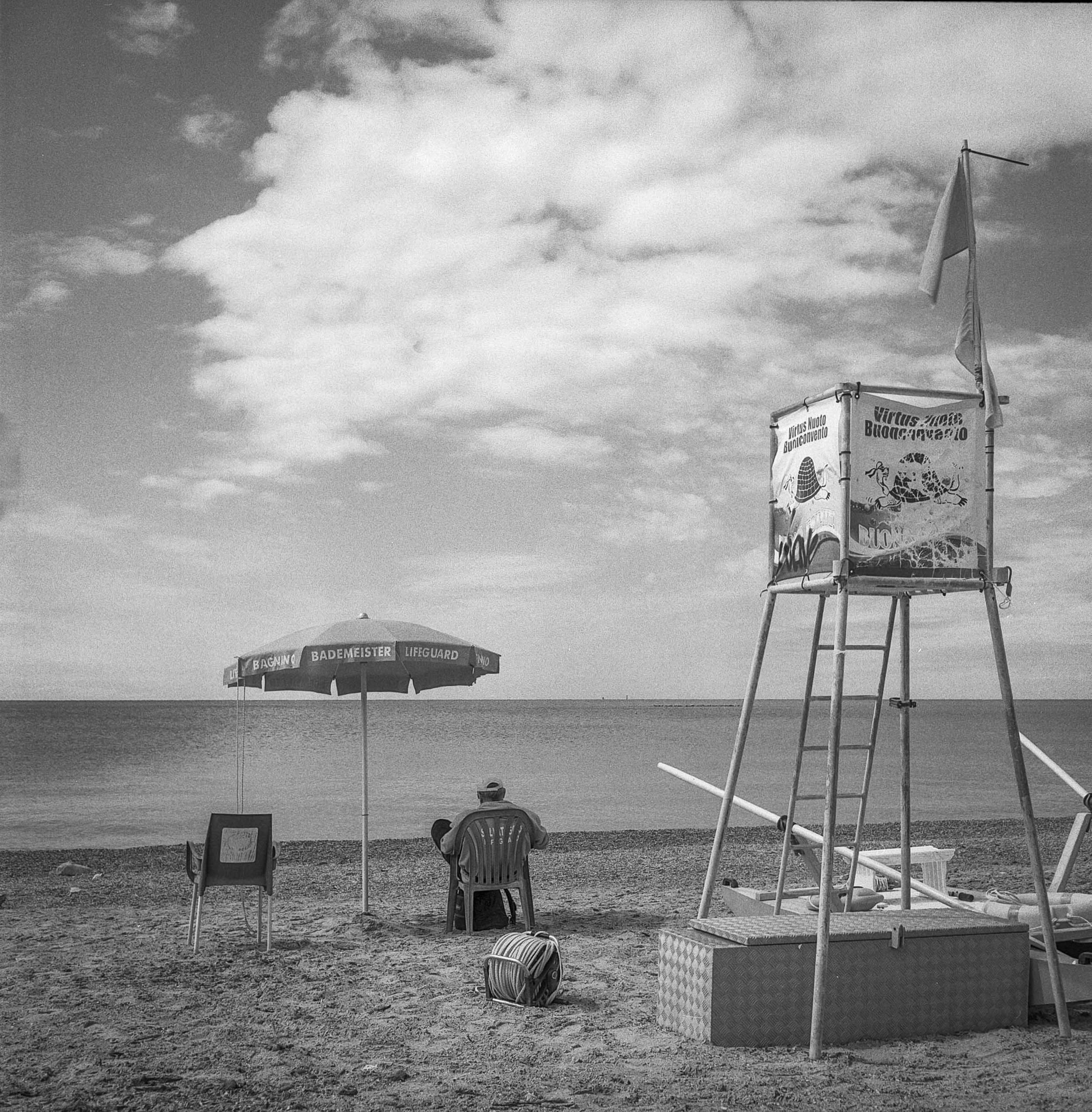 Strand mit Sonnenschirm, Strandkorb und Lifeguard-Stand mit Wimpel, bei bewölktem Himmel, schwarz-weiß Fotografie.