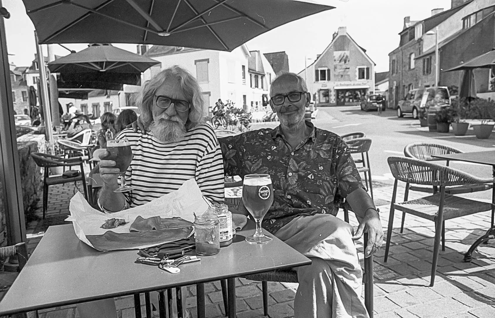 Zwei Männer sitzen an einem Tisch in einem Café im Freien, trinken Bier und lächeln. Im Hintergrund sind weitere Gäste und Häuser in einer kleinen Stadt.