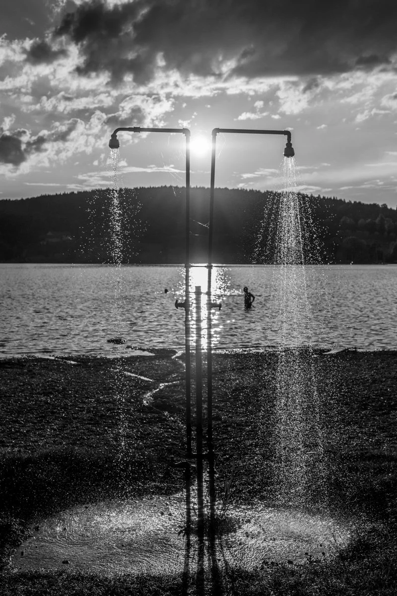 Zwei Wasserhähne mit Wasser, Sonne im Himmel auf einem See, Silhouetten von Menschen im Wasser, Berge im Hintergrund, schwarz-weiße Fotografie.