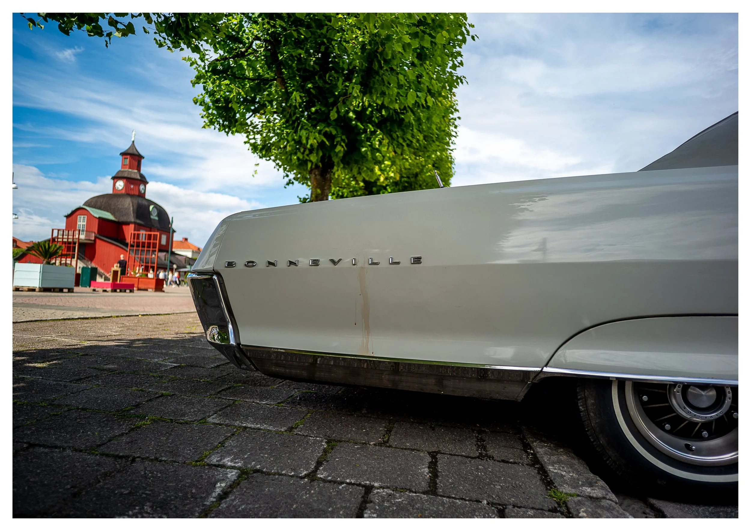 Ein alter weiße Cadillac mit dem Schriftzug 'BONNEVILLE' auf der Seite, davor ein Kopfstück eines Baumes, im Hintergrund eine rot gestrichene Kirche mit einem Uhrturm, Menschen im öffentlichen Platz, Himmel mit wenigen Wolken.