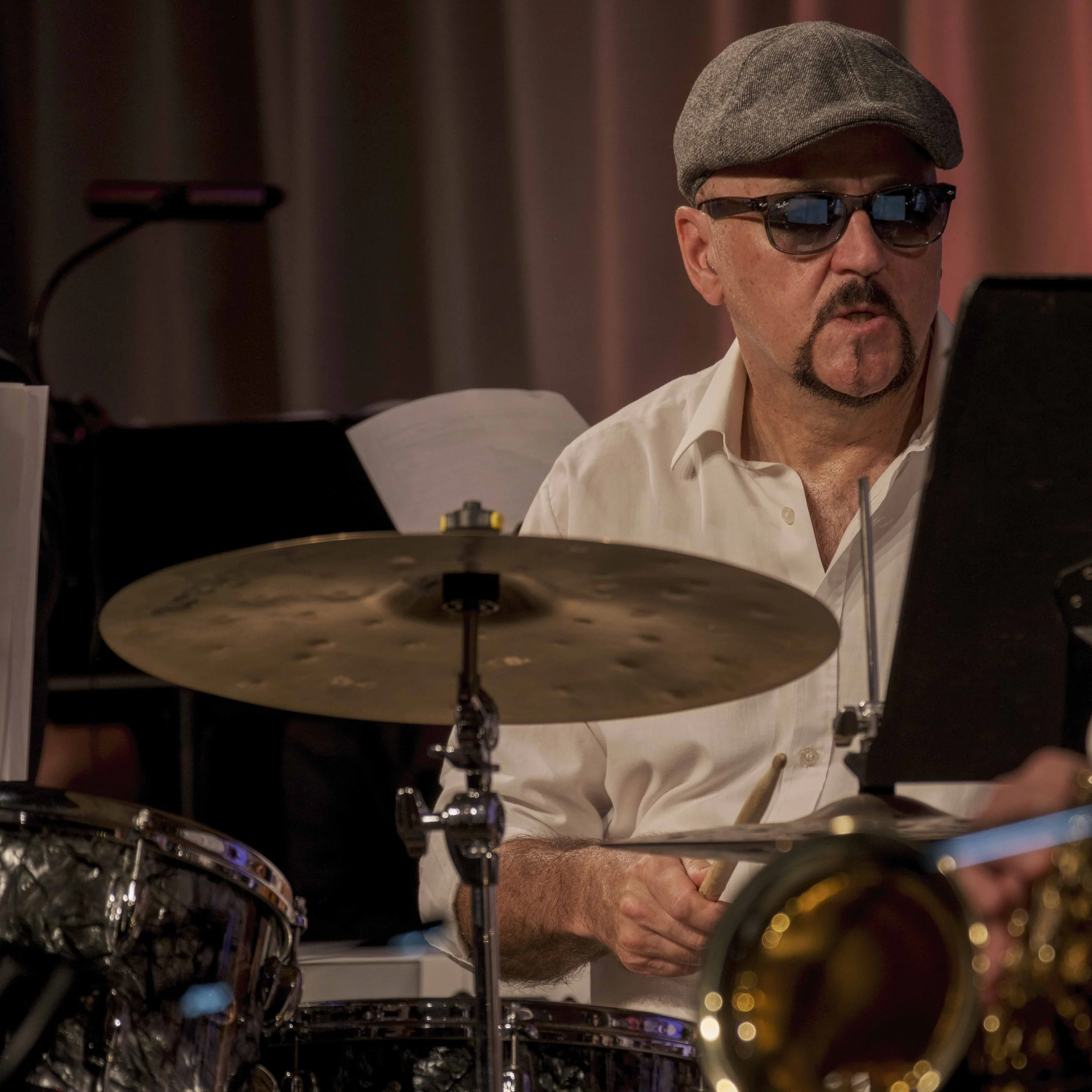 Ein Mann trägt eine graue Baskenmütze, große schwarze Sonnenbrille und ein weißes Hemd. Er sitzt hinter einem Schlagzeug, das im Vordergrund sichtbar ist, und scheint sich auf das Spielen zu konzentrieren.