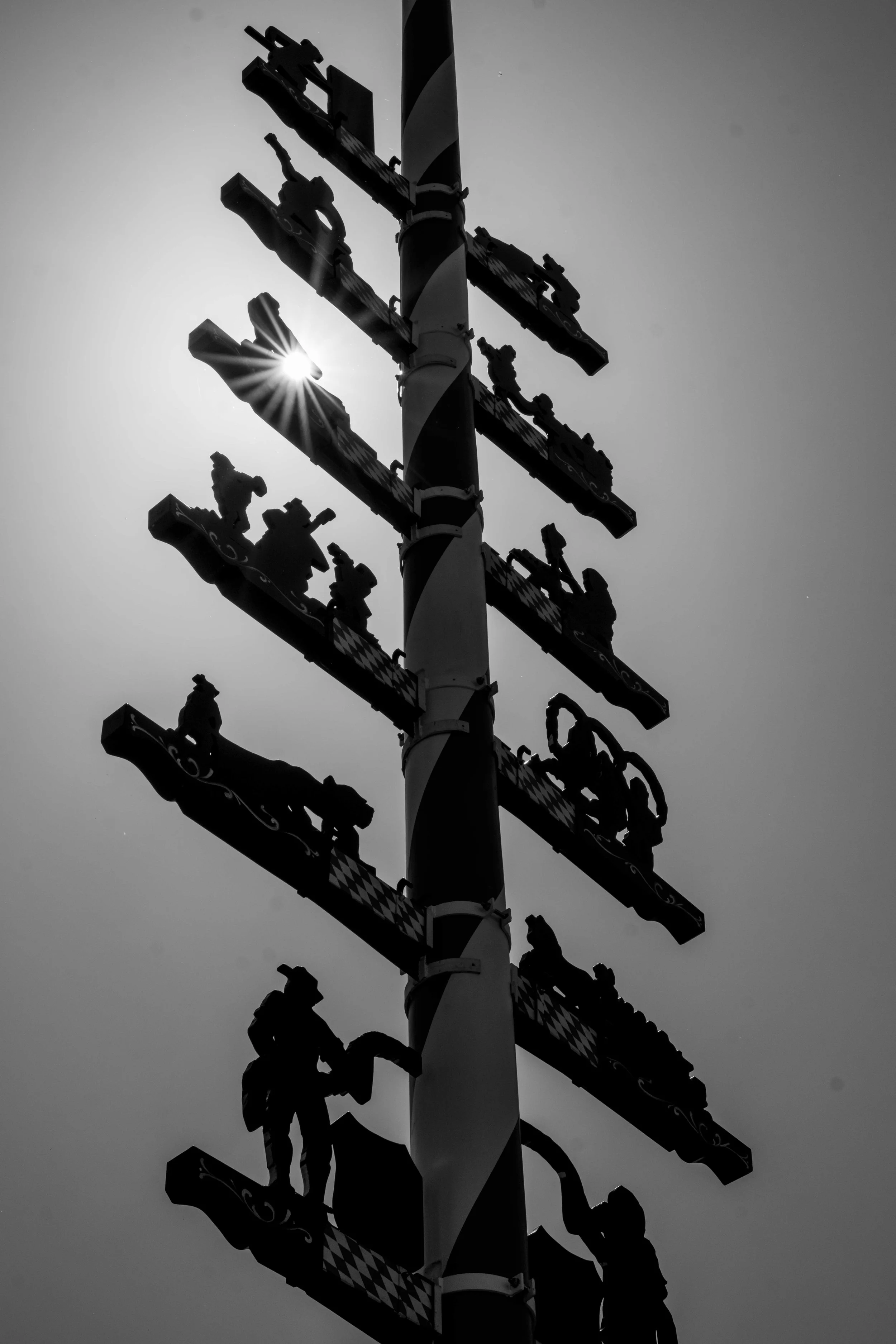 Schwarze Silhouetten von Menschen auf einer hohen, vertikal angeordneten Fahrattraktion, bei Sonnenlicht im Hintergrund, in Schwarz-Weiß