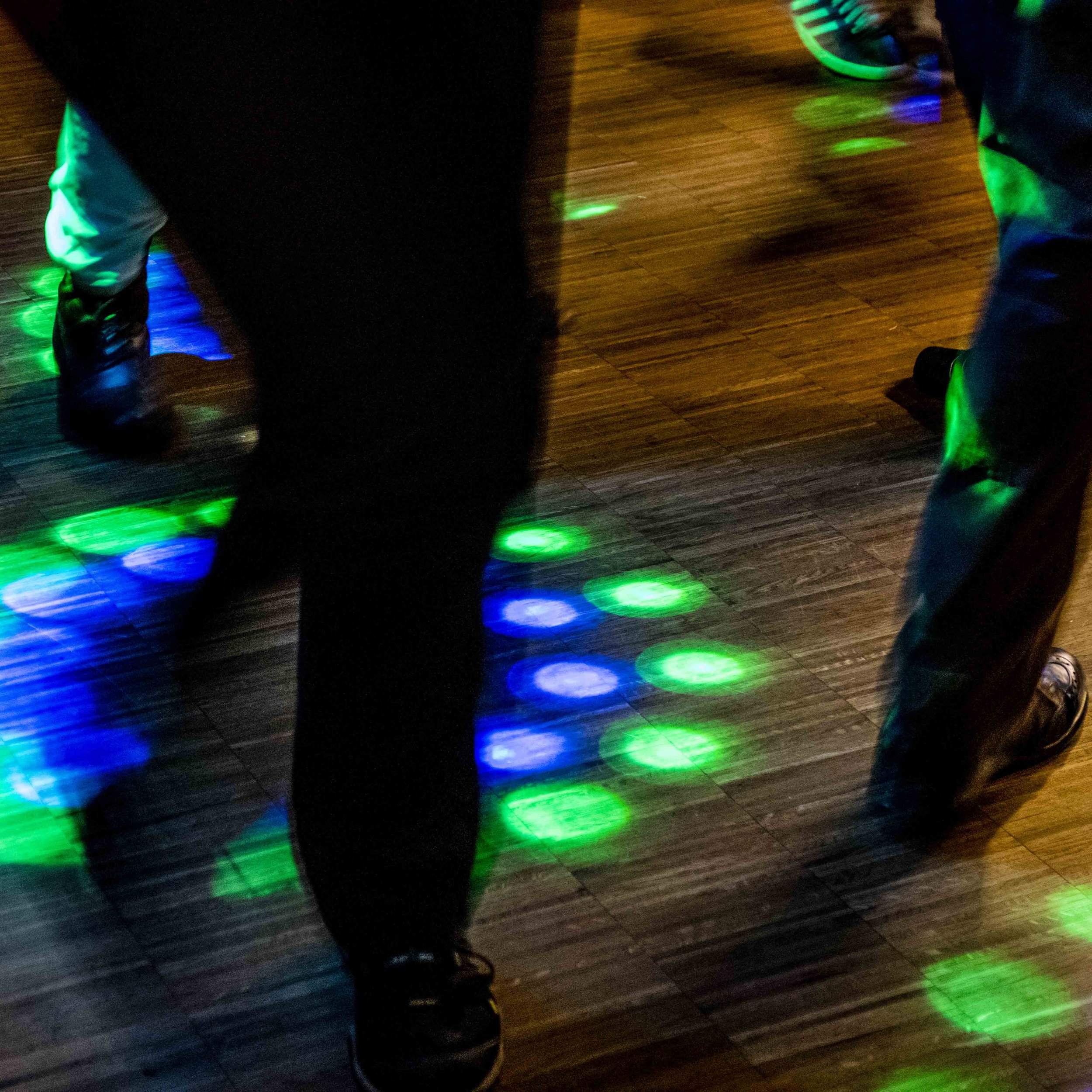 Blick auf eine Tanzfläche mit bunten Discokugeln an der Decke, auf der Tanzboden ist ein Paar mit Laufschuhen. Das Licht ist dunkel, die Farben sind lebhaft.